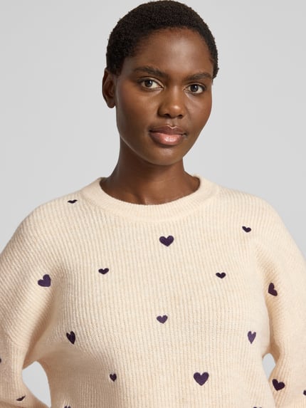 Frauenmodel trägt Strickpullover mit Woll-Anteil von Apricot in Creme mit violetten Herz-Stitchings, Ripp-Optik, Nahaufnahme Oberkörper.