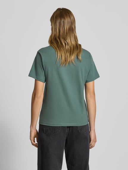 Kobiece modelka nosi T-shirt mit Brusttasche und Label-Detail od Carhartt Work In Progress z bawełny, krój regular fit, okrągły dekolt, brustasza, detal z logo, długość tyłu 59 cm w rozmiarze S, widok całej sylwetki od tyłu.