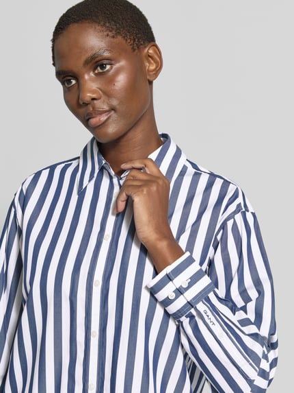 Frauenmodel trägt Relaxed Fit Langarmbluse aus reiner Baumwolle von GANT, blau-weiß gestreift, glatte Textur, Nahaufnahme Oberkörper. Damen Langarmbluse von GANT.