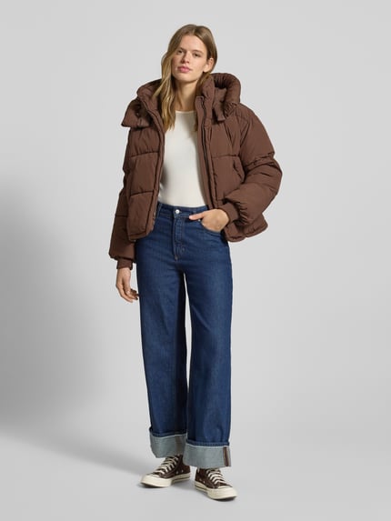 Frauenmodel trägt Regular Fit Steppjacke mit Kapuze Modell 'CORNELIA' von Only in Braun, kombiniert mit Jeans, Ganzkörperansicht von vorne.