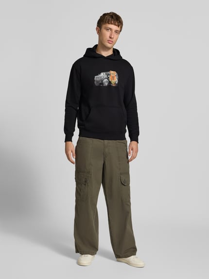 Männermodel trägt Relaxed Fit Hoodie aus Baumwoll-Mix mit Motiv-Print von CHILL PILL, schwarze Farbe, Känguru-Tasche, elastische Abschlüsse, Ganzkörperansicht von vorne.
