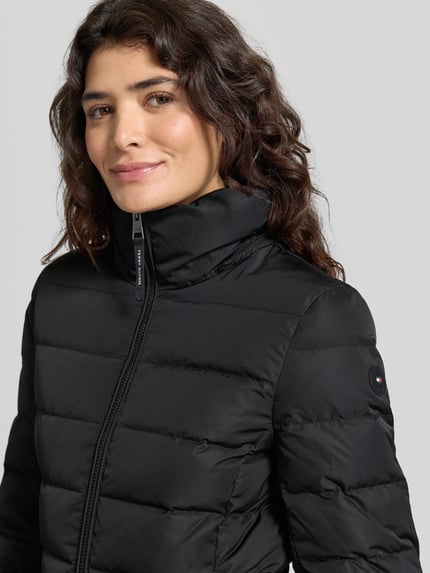 Frauenmodel trägt Daunenmantel mit Kapuze von Tommy Hilfiger in einfarbigem Schwarz mit Steppnähten, Logo-Detail, Nahaufnahme Oberkörper.