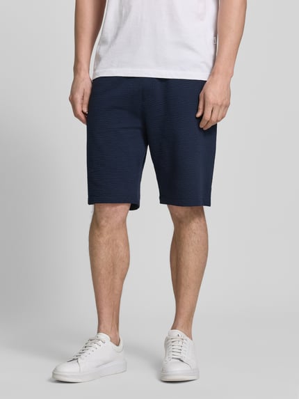 Männermodel trägt Relaxed Fit Bermudas aus Baumwoll-Mix Modell 'DETROIT' von s.Oliver RED LABEL in Dunkelblau mit Waffelstruktur, Nahaufnahme Oberkörper bis Knie, unifarbene Optik mit elastischem Bund und Eingrifftaschen.