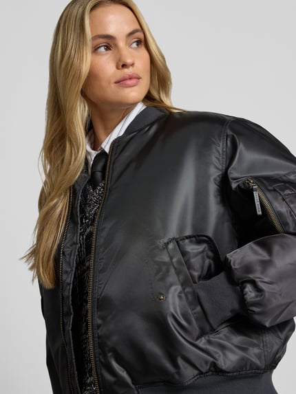 Modelka kobieca nosi Bomberjacke mit Pattentaschen od Anine Bing, czarna, luźny krój, zamek na całej długości, stójka, ściągacze, kieszenie z patką i na ramieniu, długość tyłu w rozm. S: 64 cm, szerokość w rozmiarze S: 60 cm, długość rękawa w rozmiarze S: 60 cm, zbliżenie na górną część ciała