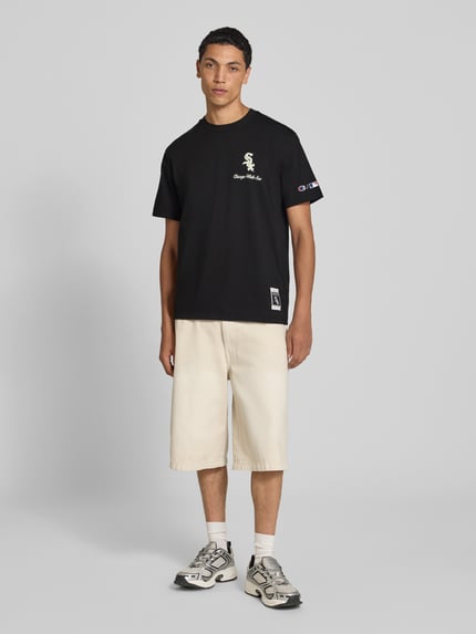 Männermodel trägt Relaxed Fit T-Shirt aus reiner Baumwolle von Champion in Schwarz mit Statement-Print, helle Shorts, Ganzkörperansicht von vorne.