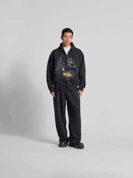 Männermodel trägt Oversized Sweatjacke mit Foto-Print von VETEMENTS aus Baumwoll-Mix mit Zweiwege-Reißverschluss, Kapuze mit Tunnelzug, dunkle Farben, Rückenlänge bei Größe M 74 cm, Schulterbreite 68 cm, Ärmellänge 67 cm, Oversized-Fit, Ganzkörperansicht von vorne.