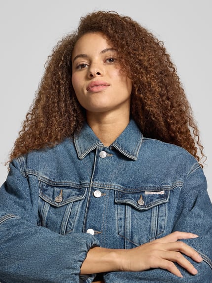Frauenmodel trägt Cropped Jeansjacke mit Knopfleiste von Calvin Klein Jeans in Blau, Reine Baumwolle, Lockerer-Schnitt, Brusttaschen, Nahaufnahme Oberkörper.