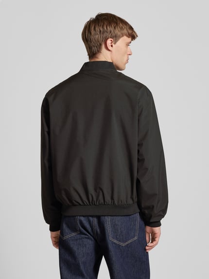 Mężczyzna nosi Bomberjacke mit seitlichen Eingrifftaschen Modell 'CHARGE' von Jack & Jones, czarna, krój straight fit, długość tyłu w rozmiarze M: 66 cm, szerokość ramion w rozmiarze M: 54 cm, szerokość klatki piersiowej w rozmiarze M: 65 cm, długość rękawa w rozmiarze M: 64 cm, widok całej sylwetki z tyłu