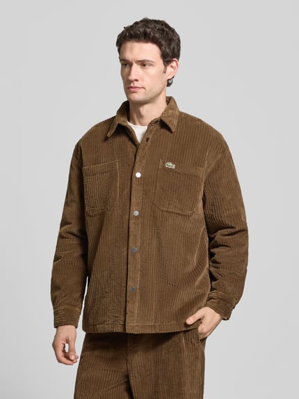 Männermodel trägt Regular Fit Cordhemd mit Logo-Patch von LACOSTE in Braun, Strukturcord, Knopfleiste, Brusttasche, unifarben, Ganzkörperansicht von vorne.