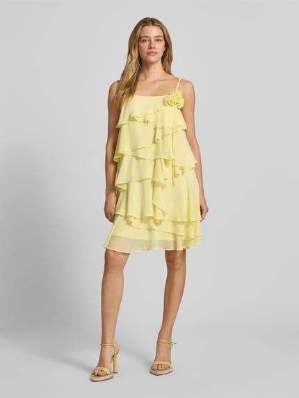 Modelka kobieca ma na sobie Fließendes Cocktailkleid mit Volants Modell 'ALADYN' od Vero Moda, jasny żółty, jednolity, krój luźny, mini, dekolt kwadratowy, ozdobny kwiat, falbany, regulowane cienkie ramiączka, widok całej sylwetki z przodu.