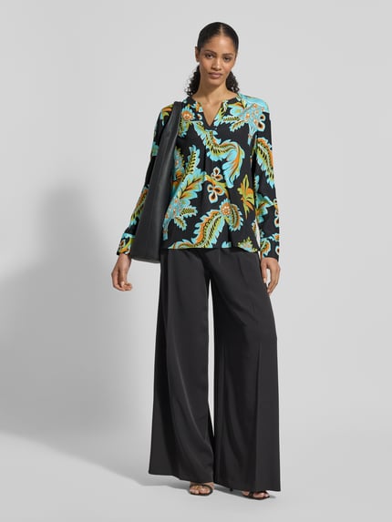 Modelka nosi Regular Fit Bluse aus Seide-Elasthan-Mix von HERZENS o wzorze allover, tunikowy kołnierz, czarne szerokie spodnie, ujęcie całopostaciowe z przodu.