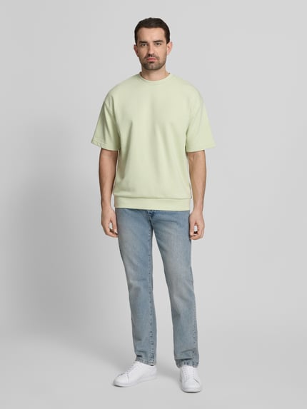 Männermodel trägt Oversized T-Shirt mit Rundhalsausschnitt von Lindbergh aus Baumwoll-Mix mit Label-Detail in Hellgelb, Ganzkörperansicht von vorne. Rückenlänge bei Größe M: 70 cm.