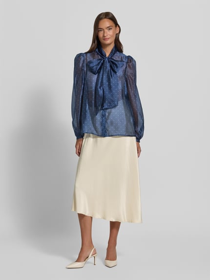 Frauenmodel trägt Schluppenbluse im semitransparenten Design von Rich & Royal in Blau, gepunktet, kombiniert mit cremefarbenem Rock, Ganzkörperansicht von vorne. Gerader Schnitt, Schluppe, verdeckte Knopfleiste, Rückenlänge 64 cm, Ärmellänge 66 cm.