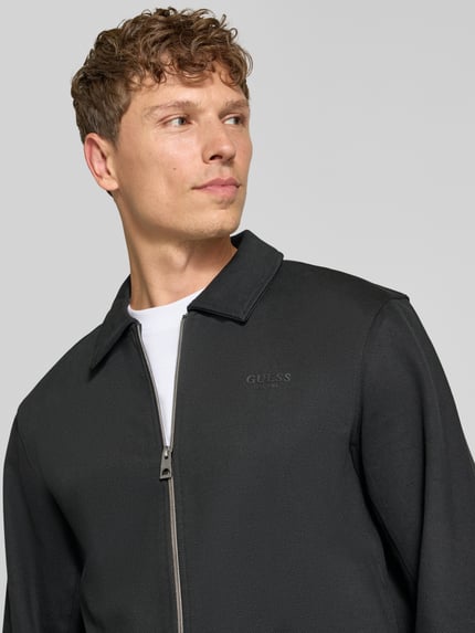Männermodel trägt Regular Fit Sweatjacke aus elastischem Baumwoll-Mix von GUESS in Schwarz mit Umlegekragen, Nahaufnahme Oberkörper.