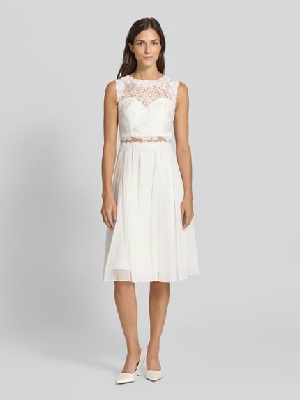 Frauenmodel trägt Cocktailkleid mit Spitze und Rundhalsausschnitt von Mascara, weißer Chiffon, A-Linie, Midikleid, Ganzkörperansicht von vorne.