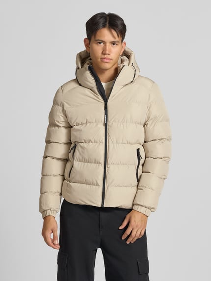 Männermodel trägt Steppjacke mit Label-Stitching und Kapuze von Superdry in Beige, seitliche Eingrifftaschen, Ganzkörperansicht von vorne.