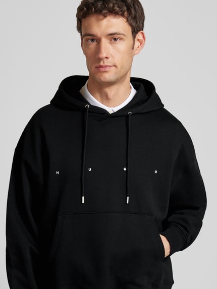 Męski model nosi Relaxed Fit Hoodie aus reiner Baumwolle Modell 'DOLAND' od HUGO, czarny, gładki, bawełna, logo, zbliżenie górnej części.