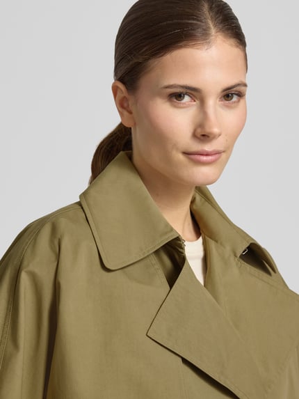 Frauenmodel trägt Trenchcoat mit verdecktem Verschluss und Bindegürtel von Blonde No. 8 in Olivgrün, Baumwoll-Mix, Nahaufnahme Oberkörper.