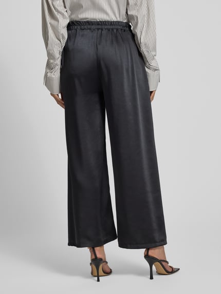Frauenmodel trägt Wide Leg Stoffhose mit elastischem Bund von ZABAIONE in Schwarz, lockere Form, leicht glänzende Textur, Ganzkörperansicht Rückansicht. Bundweite bei Größe S 34 cm, Innenbeinlänge 72 cm.