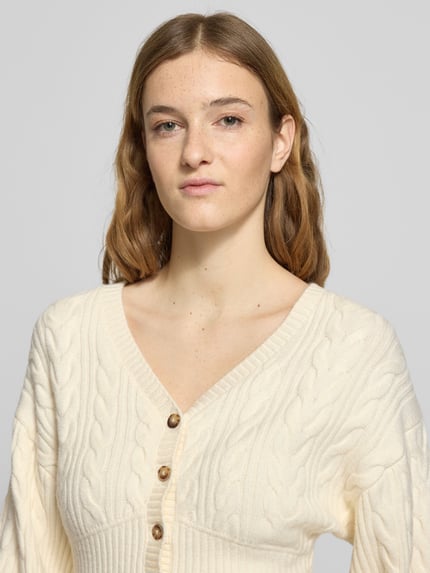 Vrouwenmodel draagt Strickjacke mit Knopfleiste van Gina Tricot in crèmekleur, viscosemix, regular fit, geribde boorden, V-hals, knoopsluiting, close-up bovenlichaam.