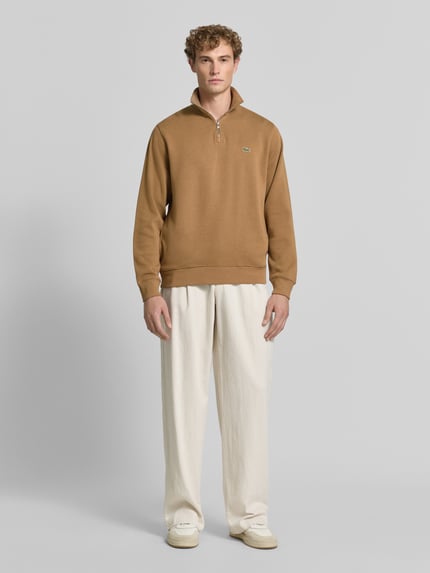 Männermodel trägt Regular Fit Troyer aus reiner Baumwolle von Lacoste in Braun, kombiniert mit heller Hose, Ganzkörperansicht von vorne. Herren Troyer von Lacoste