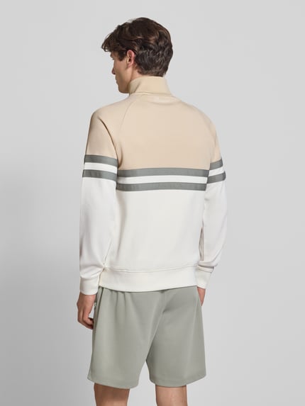 Männermodel trägt kurtka treningowa z mieszanki bawełny ellesse Trainingsjacke mit Label-Patch und Stehkragen Modell 'RIMINI', krój regular fit, wpuszczane kieszenie, zamek błyskawiczny, stójka, długość tyłu w rozmiarze M: 71 cm, długość rękawów w rozmiarze M: 65 cm, kolor beżowo-biały z szarymi paskami, gładka tekstura, Ganzkörperansicht Rückansicht.