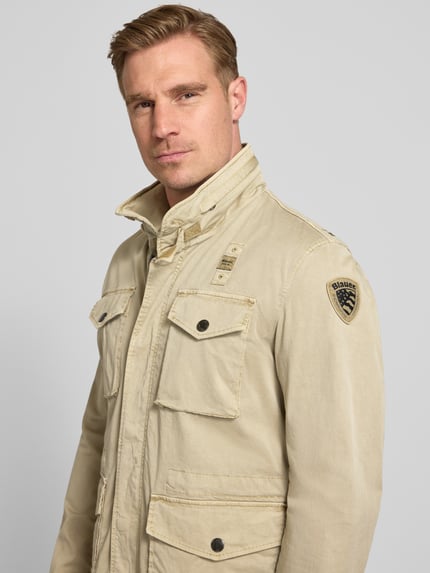 Männermodel trägt heren jack Jacke mit Stehkragen und Pattentaschen Modell 'Ashmont' van Blauer Usa, mix van katoen en elastaan, regular fit, vier klepzakken, knopen en badge op de mouw, beige kleur, gladde textuur, halflichaamaanzicht van voren. Ruglengte maat M: 72 cm, schouderbreedte: 40 cm, borstwijdte: 52 cm, mouwlengte: 63 cm.