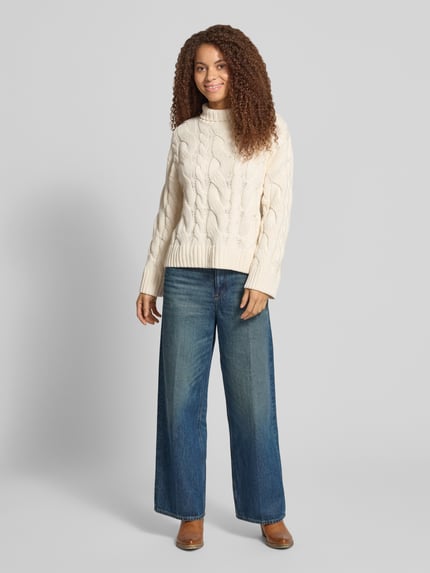 Frauenmodel trägt Regular Fit Strickpullover aus Baumwoll-Mix von Gina Tricot, cremefarbenes Zopfmuster, Stehkragen, Jeans, Ganzkörperansicht von vorne.