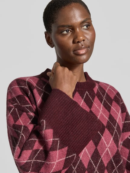 Kobiece modelka nosi Relaxed Fit Pullover mit Woll-Anteil od Neo Noir w elastycznej mieszance materiałów z dodatkiem wełny, imitacja dzianiny, okrągły dekolt, wzór w odcieniach bordowego i różowego, długość tyłu w rozmiarze 36: 54 cm, długość rękawów w rozmiarze 36: 50 cm, zbliżenie na górną część ciała