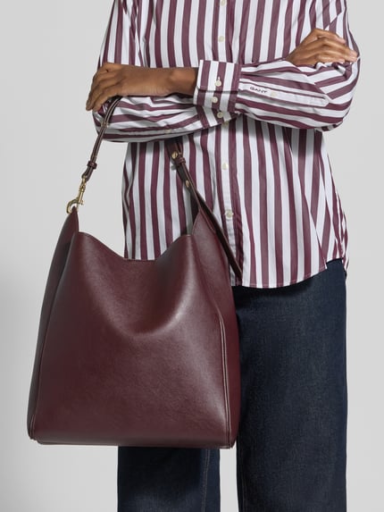 Frauenmodel trägt Handtasche mit Magnetverschluss Modell 'DALMA' von MANGO in Dunkelrot, glatte Textur, Nahaufnahme Oberkörper. Hauptfach mit Magnetverschluss, verstellbarer Schulterriemen, Karabinerverschluss, Maße: 31x33x11 cm, Schulterriemen 52 cm.