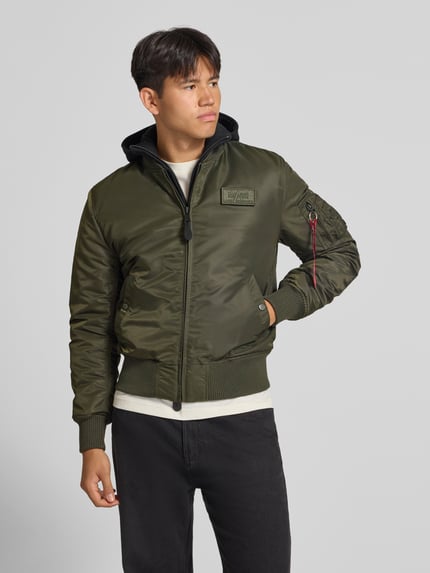 Männermodel trägt Jacke mit Reißverschlusstasche am Ärmel von Alpha Industries in Olivgrün mit Label-Patch, Ganzkörperansicht von vorne. Regular Fit, Kapuze mit Reißverschluss, gerippter Stehkragen, Reißverschluss, Eingrifftaschen mit Druckknopfverschluss, gerippte Abschlüsse, Rückenlänge bei Größe M 64 cm, Schulterbreite 54 cm, Brustweite 54 cm, Ärmellänge 72 cm.