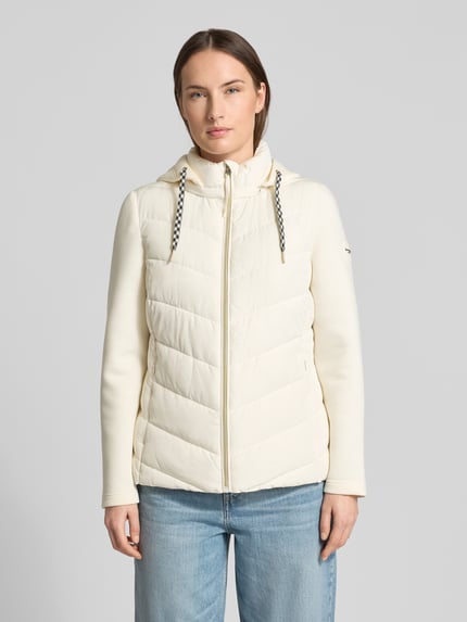 Modelka kobieca nosi Slim Fit Steppjacke mit gefüttertem Stehkragen von Christian Berg Woman Selection, kremowy kolor, krój slim fit, kaptur z kołnierzem, zamek błyskawiczny, kieszenie na zamek, długość pleców w rozmiarze 36: 60 cm, szerokość ramion w rozmiarze 36: 41 cm, szerokość klatki piersiowej w rozmiarze 36: 51 cm, długość rękawa w rozmiarze 36: 62 cm; widok całej sylwetki od przodu.