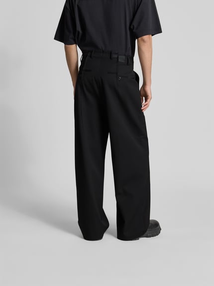 Männermodel trägt schwarze Straight Fit Hose aus Schurwoll-Mix von VETEMENTS mit Bundfalten und Gesäßtaschen, Ganzkörperansicht Rückansicht.