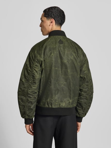 Männermodel trägt Bomberjacke mit Label-Print von Don’t Waste Culture, krój regular fit, stójka, zamek błyskawiczny dwustronny, elastyczne wykończenia, nadruk z logo, długość tyłu w rozmiarze M: 67 cm, szerokość w rozmiarze M: 53 cm, długość rękawów w rozmiarze M: 64 cm, odcienie zieleni, lekko połyskująca tekstura, czarne ściągacze, całkowity widok z tyłu.