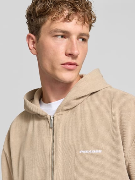 Männermodel trägt Oversized Fit Sweatjacke mit Label-Detail Modell 'Mezzano' von PEGADOR in Beige, Material-Mix, Nahaufnahme Oberkörper.