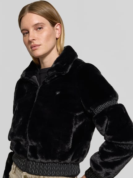 Vrouwenmodel draagt Jacke aus Kunstfell mit Stehkragen Modell 'CARLA' van GUESS Activewear, zwart kunstbont met glanzende textuur, regular fit, details aan boord en mouwen, ritssluiting, borstbreedte 50 cm bij maat S, schouderbreedte 37 cm, ruglengte 49 cm en mouwlengte 59 cm, close-up bovenlichaam.