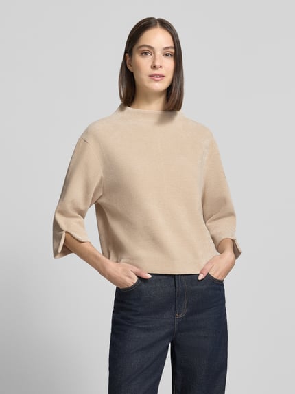 Frauenmodel trägt Relaxed Fit Strickpullover mit Stehkragen von ZABAIONE in Beige, Strick-Optik, 3/4-Arm, Logo-Emblem, Ganzkörperansicht von vorne.