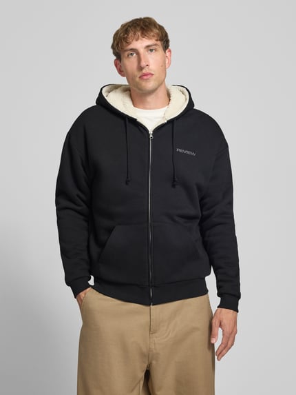 Männermodel trägt Regular Fit Sweatjacke aus Baumwoll-Mix mit Teddyfutter von REVIEW in Schwarz, Logo-Detail, Nahaufnahme Oberkörper.
