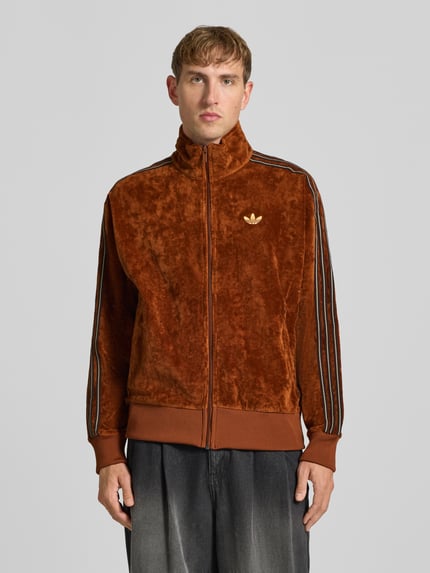 Männermodel trägt Sweatjacke mit Samtoptik Modell 'FIREBIRD TT VEL' von adidas Originals in Braun, unifarben, glänzend, Logo-Print, Ganzkörperansicht von vorne.