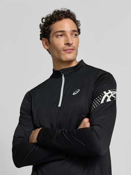 Męski model nosi bluzkę z długim rękawem Langarmshirt mit Troyer-Kragen Modell 'ICON' von ASICS TIGER, czarny z nadrukiem z logo, prosty krój, kołnierz zapinany, Długość tyłu w rozmiarze M: 72 cm, ganzkörperansicht von vorne