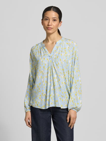 Modelka nosi Regular Fit Bluse aus reiner Viskose mit Allover-Motiv-Print od Emily Van den Bergh, jasnoniebieska z żółtym wzorem, długi rękaw, tunikowy dekolt, elastanowe mankiety, krój regular fit, widok na cały korpus z przodu.