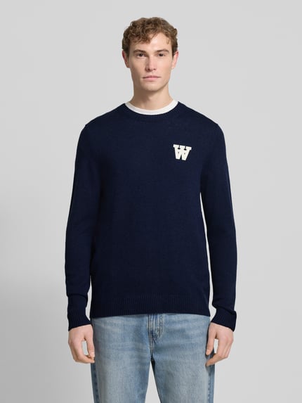 Männermodel trägt Strickpullover aus Lammwolle Modell 'Tay' von Wood Wood in Dunkelblau mit Label-Stitching, Regular-Fit, Rundhalsausschnitt, Ganzkörperansicht von vorne.