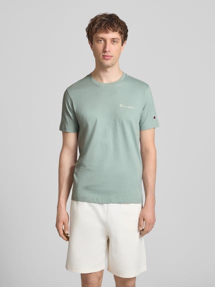 Männermodel trägt T-Shirt mit Label-Stitchings und Rundhalsausschnitt von CHAMPION in Grün, reine Baumwolle, Regular Fit, Ganzkörperansicht von vorne.