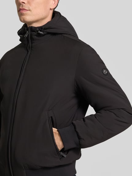 Männermodel trägt Funktionsjacke mit Kapuze Modell 'C-Caniol' von BOSS in einfarbigem Schwarz mit Reißverschlusstaschen und strukturierter Webware, Nahaufnahme Oberkörper.