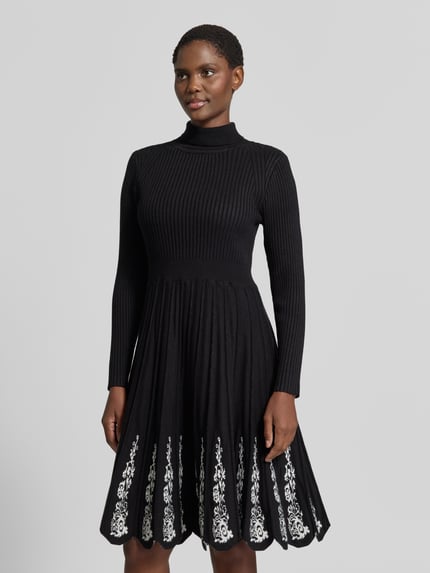 Frauenmodel trägt Knielanges Strickkleid aus Viskose-Mix von Apricot in Schwarz mit floralen Stickereien, Ripp-Optik, Stehkragen, A-Linien-Rock, taillierte Passform, langärmlig, Ganzkörperansicht von vorne.
