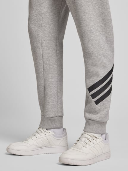 Männermodel trägt Slim Fit Sweathose mit Logo-Print von adidas Sportswear, grauer Material-Mix, elastische Beinabschlüsse, Nahaufnahme Beinbereich.