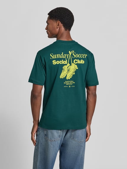 Männermodel trägt T-Shirt mit Statement-Print von MCNEAL in Dunkelgrün mit gelbem Aufdruck, lockerer Schnitt, Ganzkörperansicht Rückansicht, Rückenlänge bei Größe M 66 cm, Rundhalsausschnitt
