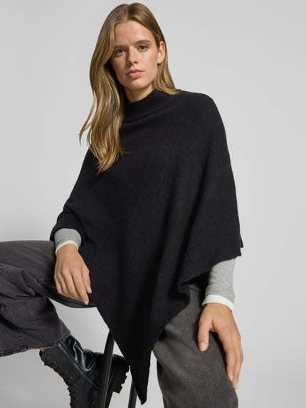 Frauenmodel trägt Poncho mit asymmetrischem Saum von PIECES in Schwarz, unifarben, Mock Neck, hochgeschlossen, Strick, Ganzkörperansicht von vorne.