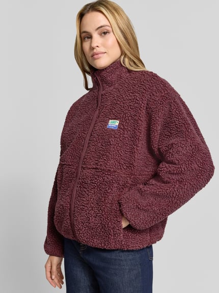 Frauenmodel trägt bordeauxrote Regular Fit Jacke aus Teddyfell Modell 'Hoktown' von American Vintage, mit Stehkragen und Logo-Patch, Nahaufnahme Oberkörper. Rückenlänge 63 cm, Schulterbreite 52 cm, Brustweite 51 cm, Ärmellänge 54 cm.