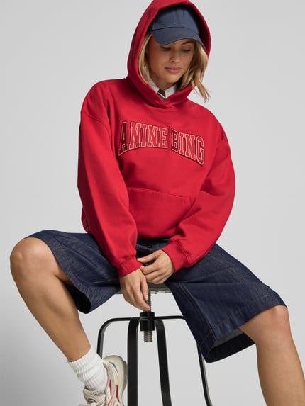 Vrouwenmodel draagt Oversized Hoodie aus reiner Baumwolle van Anine Bing in rood met labelprint, kangoeroezak, geribde boorden, hele lichaam vooraanzicht.